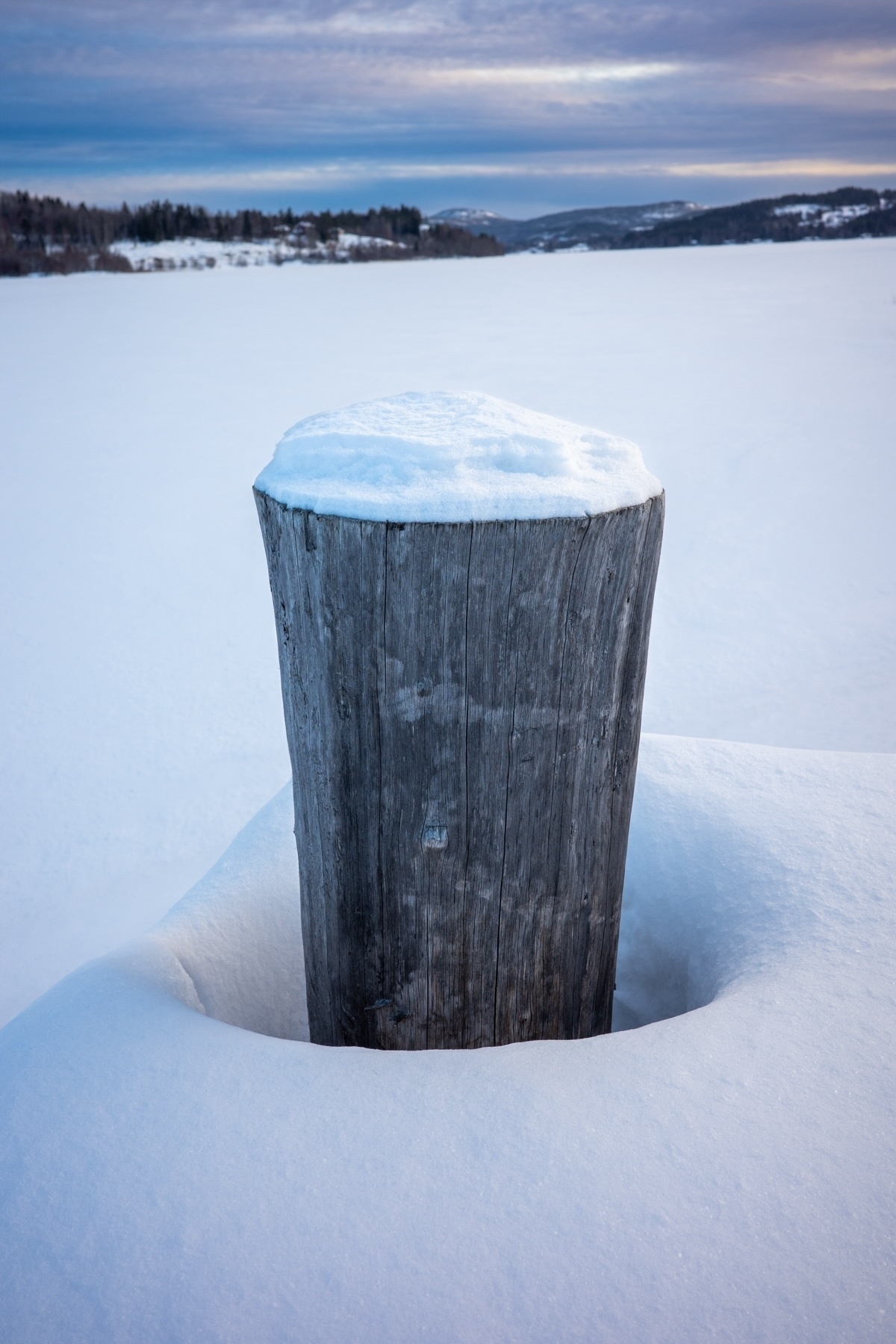 A wooden post with a cap of snow stands in the foreground against a wintery landscape with snow-covered ground and a backdrop of trees and hills under a cloudy sky.