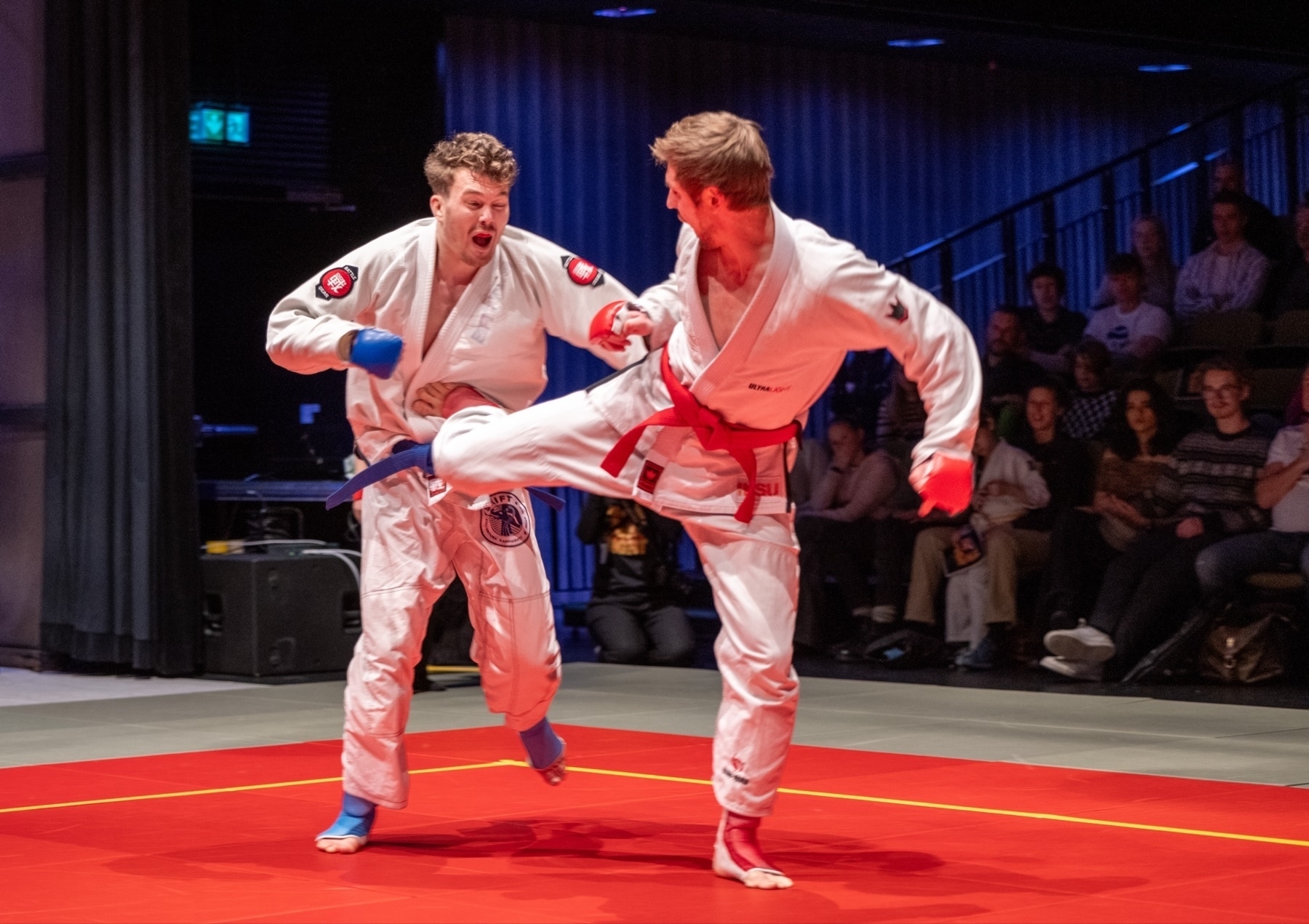 Two ju-jutsu practitioners sparring in a competition on a red mat, with one delivering a kick and the other recoiling. Spectators are visible in the background.