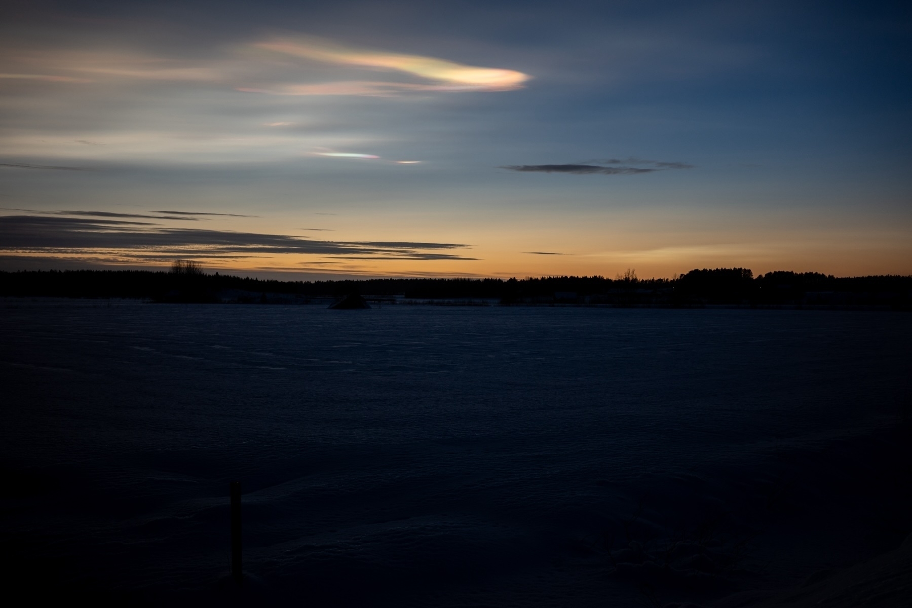 A snow-covered landscape at dusk with a colorful sky featuring a prominent streak of iridescent cloud.