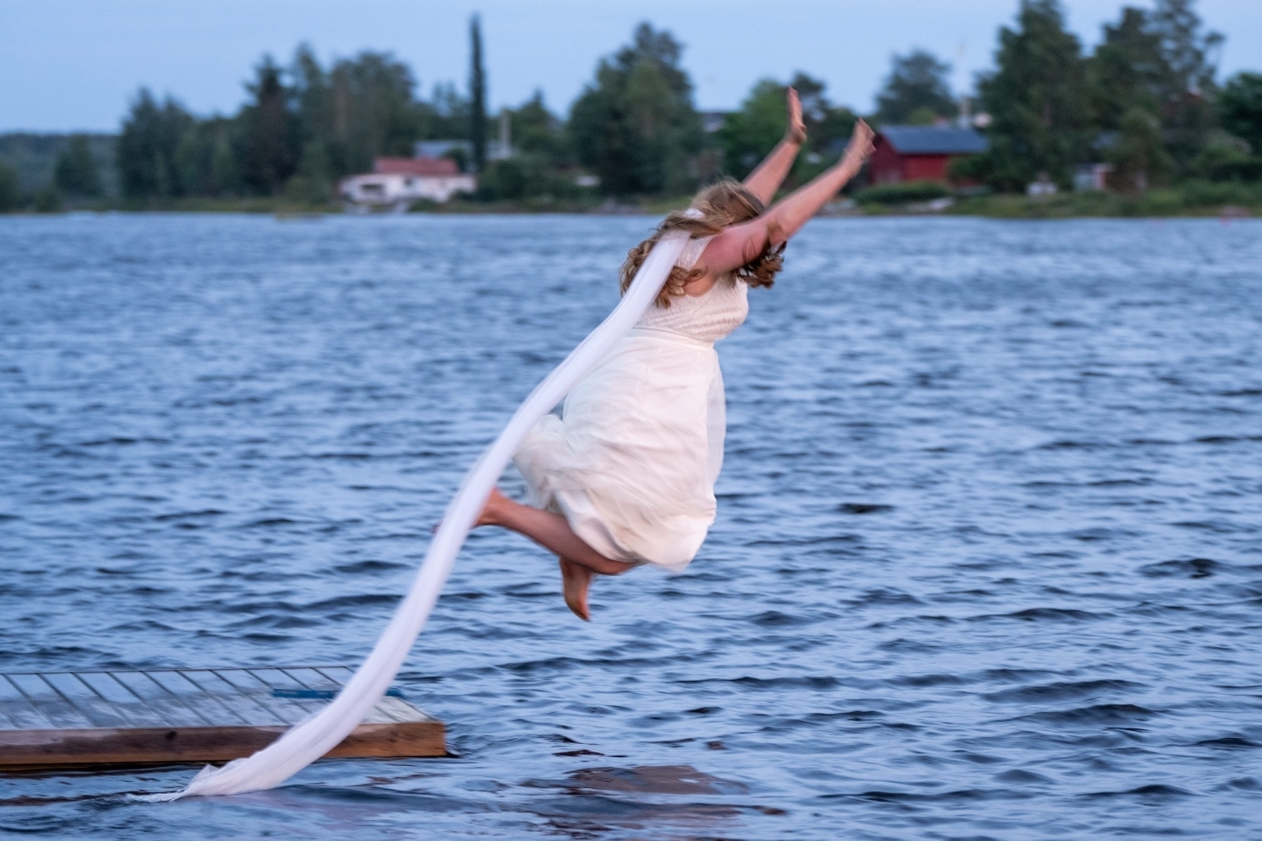 A person in a white dress jumps off a wooden platform into a lake, with a long piece of fabric trailing behind. Trees and houses are visible in the background.