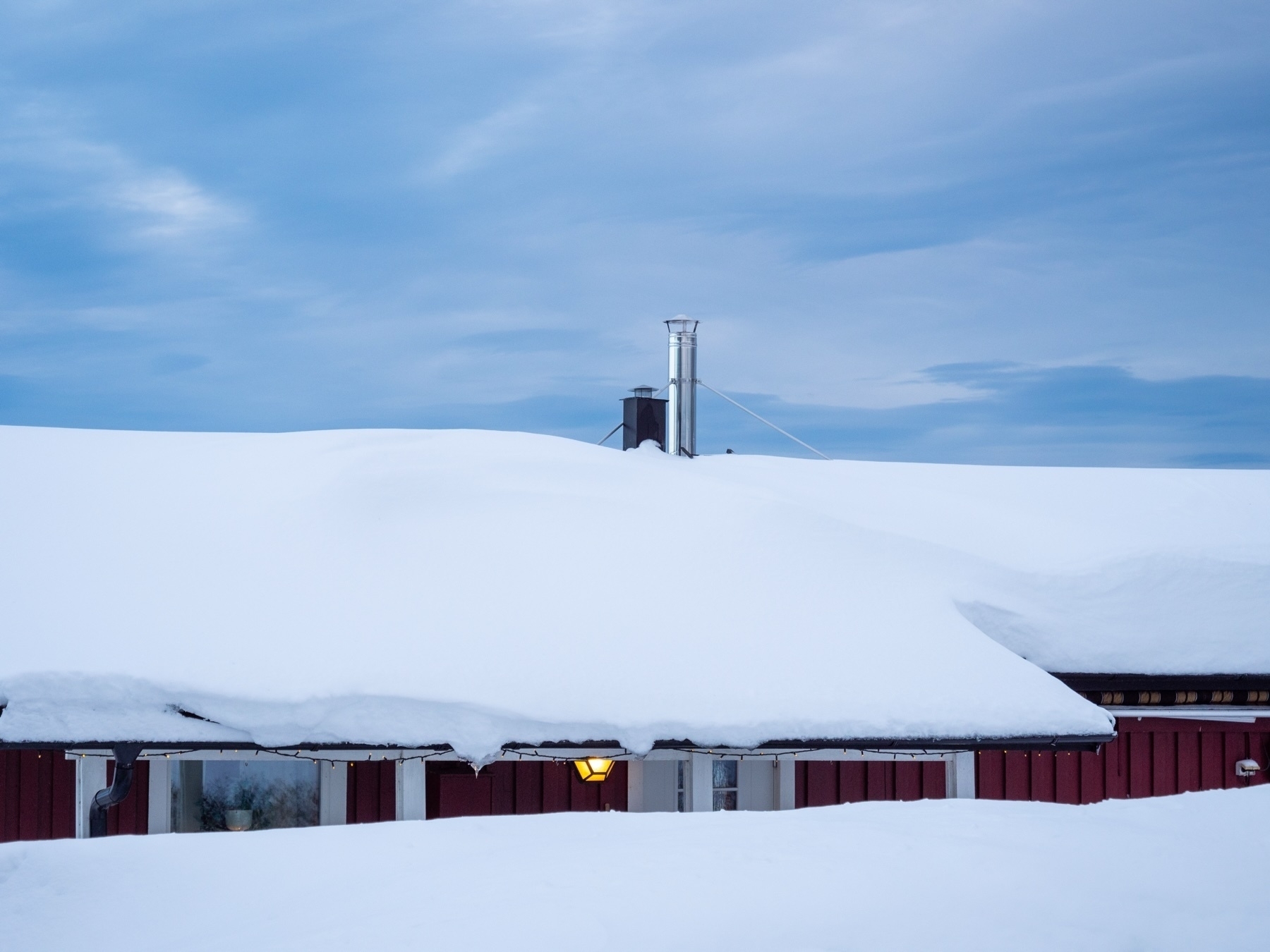 A snow-covered roof of a red building with a chimney protruding, under a cloudy blue sky.