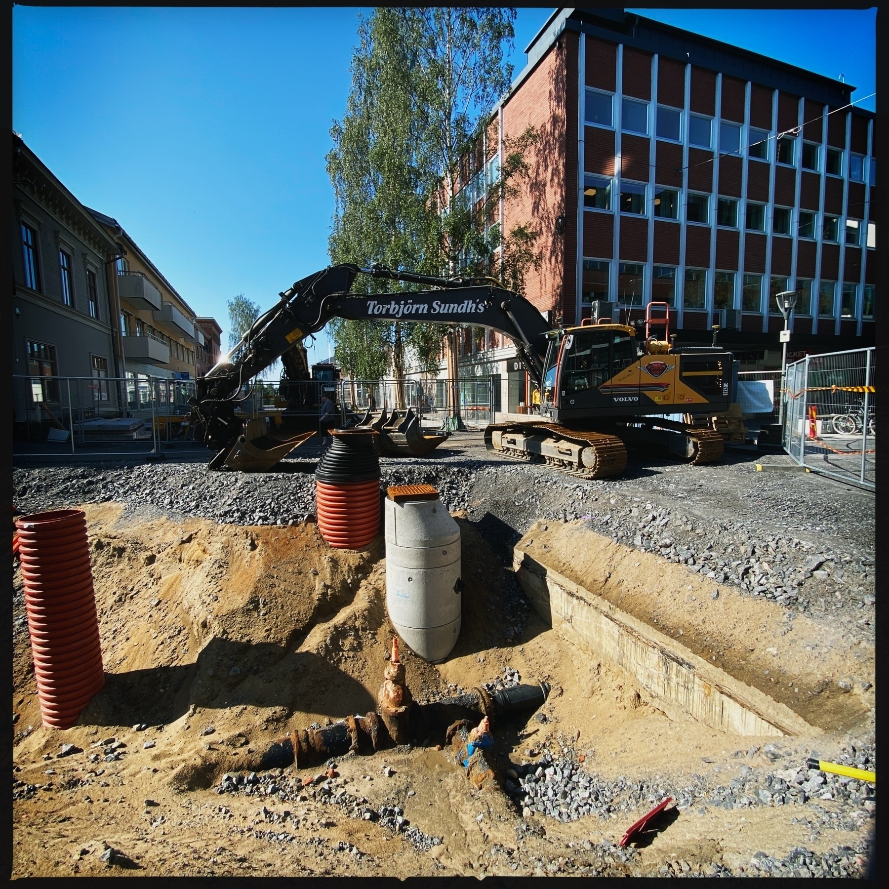 A construction site with excavation work underway. An excavator with “Torbjörn Sundh’s” written on its arm is visible, alongside construction materials such as large pipes and gravel. Surrounding the site are buildings and tall trees, with safety