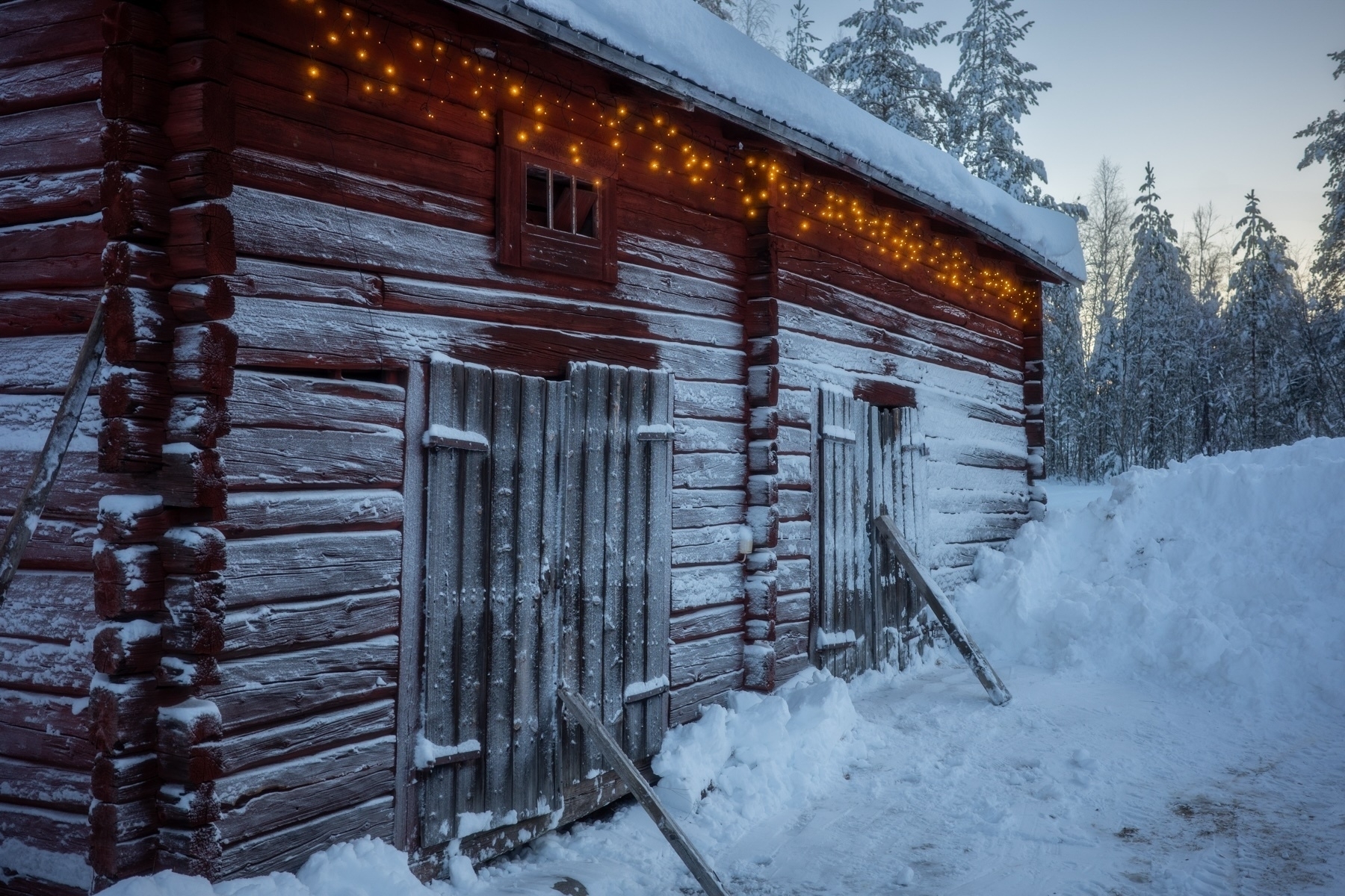 A rustic wooden cabin covered in snow with warm, glowing string lights along the eaves. The scene is set in a snowy forest landscape.