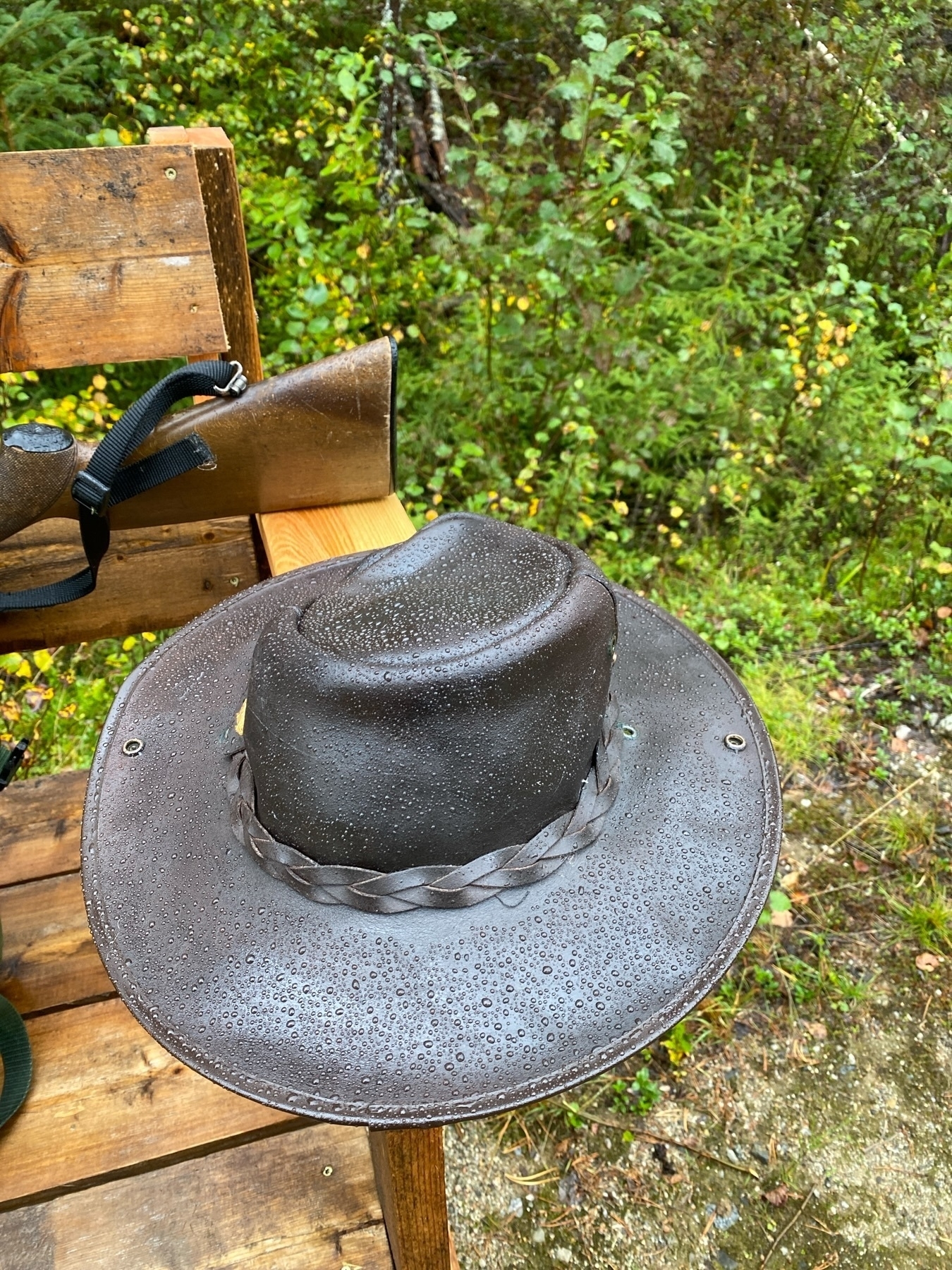 A brown leather hat covered in water droplets rests on a wooden bench outdoors, surrounded by green foliage. A strap and part of a wooden stock, possibly from a rifle, are visible behind the hat.