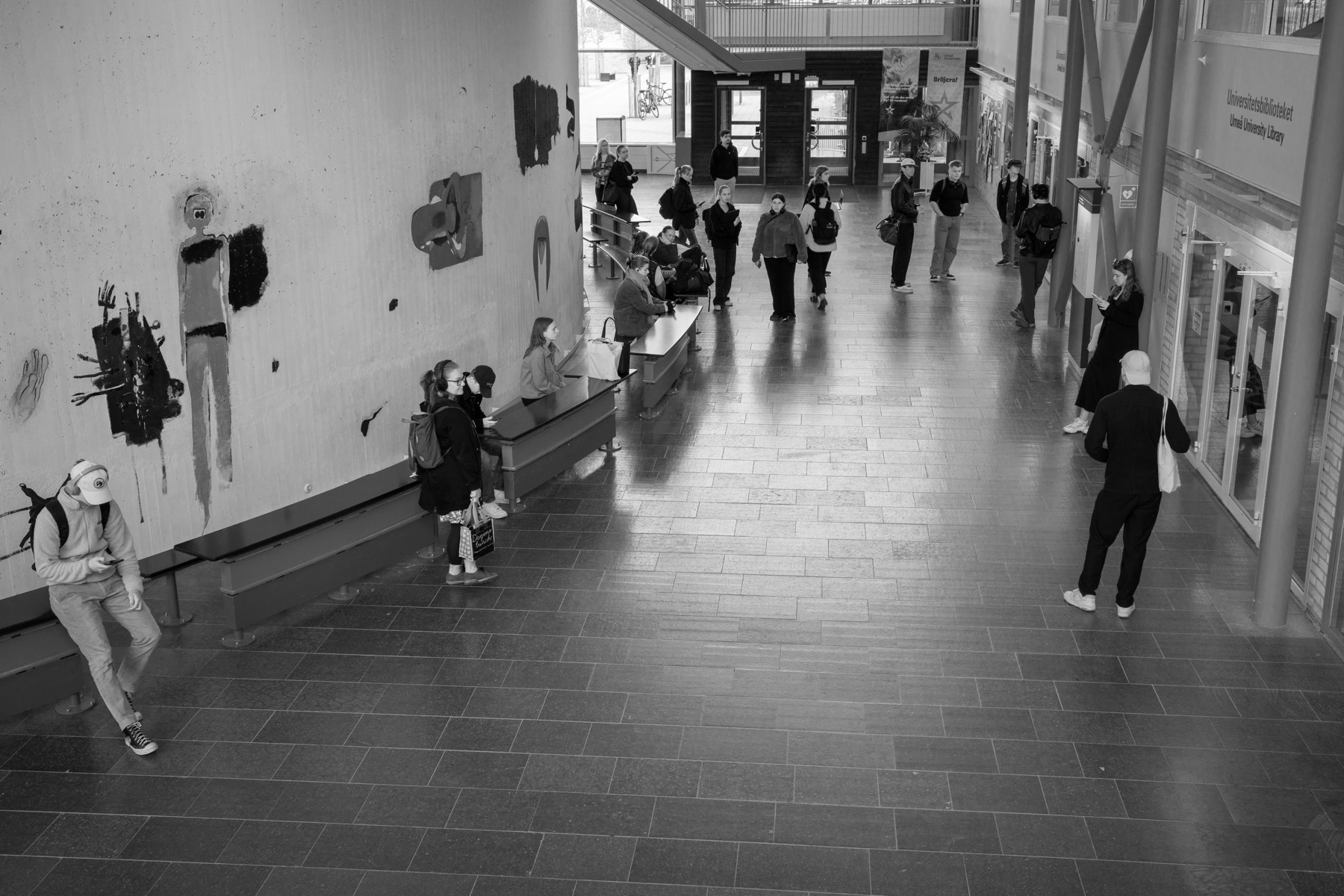A black-and-white photo of an indoor public space with several people. Some are walking, while others are sitting on benches along the wall or standing. The wall features abstract and figure paintings. In the background, there is a staircase.