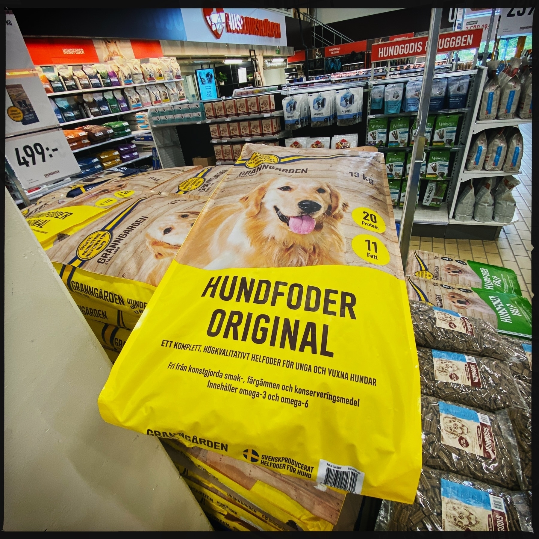 The image shows a display of yellow bags of dog food labeled “Hundfoder Original” in a pet store. The packaging features a picture of a Golden Retriever and highlights the product’s attributes, such as protein and fat content. The background includes shelves