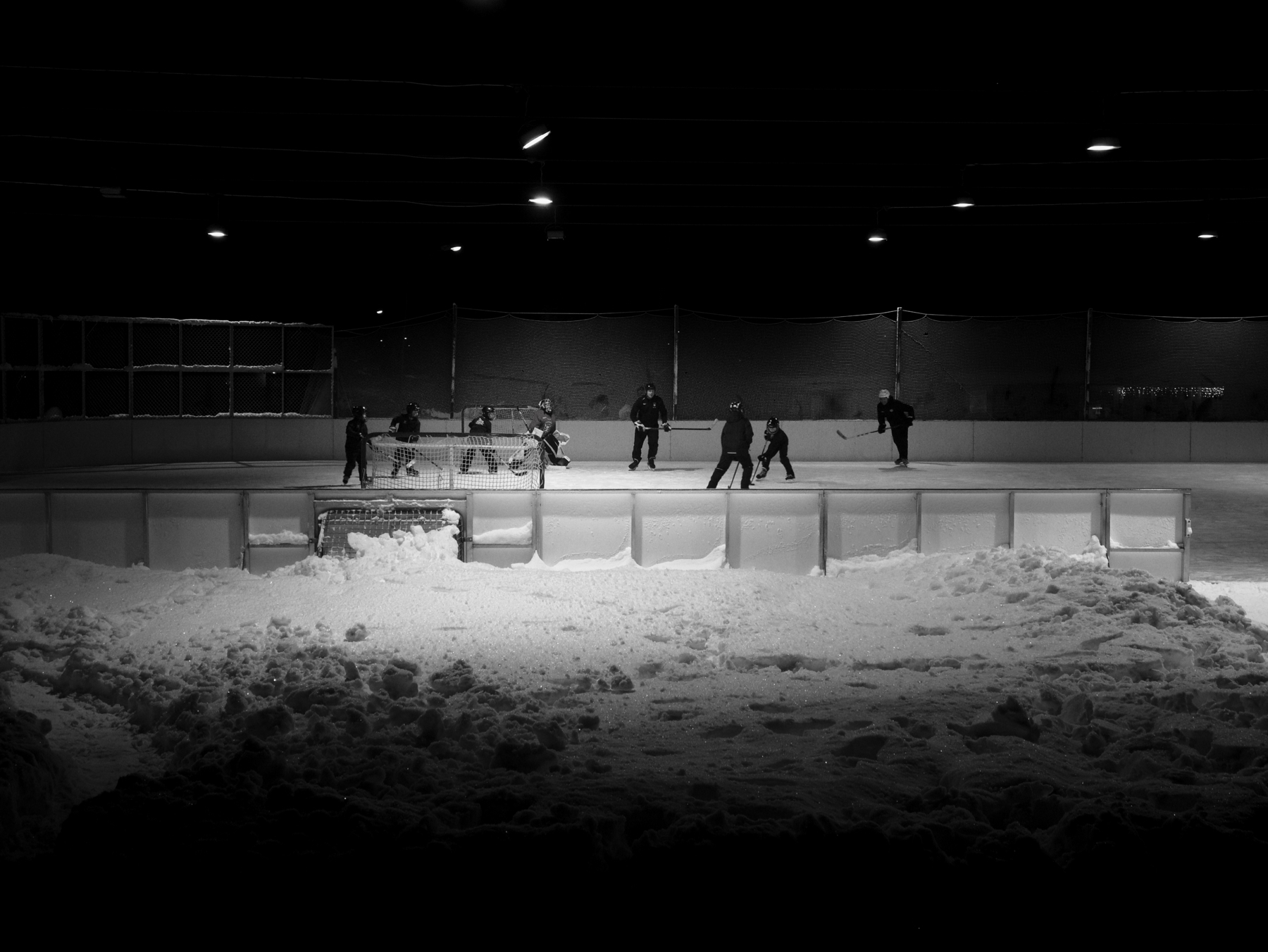 A black and white photo of people playing ice hockey on an outdoor rink at night. Snow is piled around the rink, and overhead lights illuminate the scene.