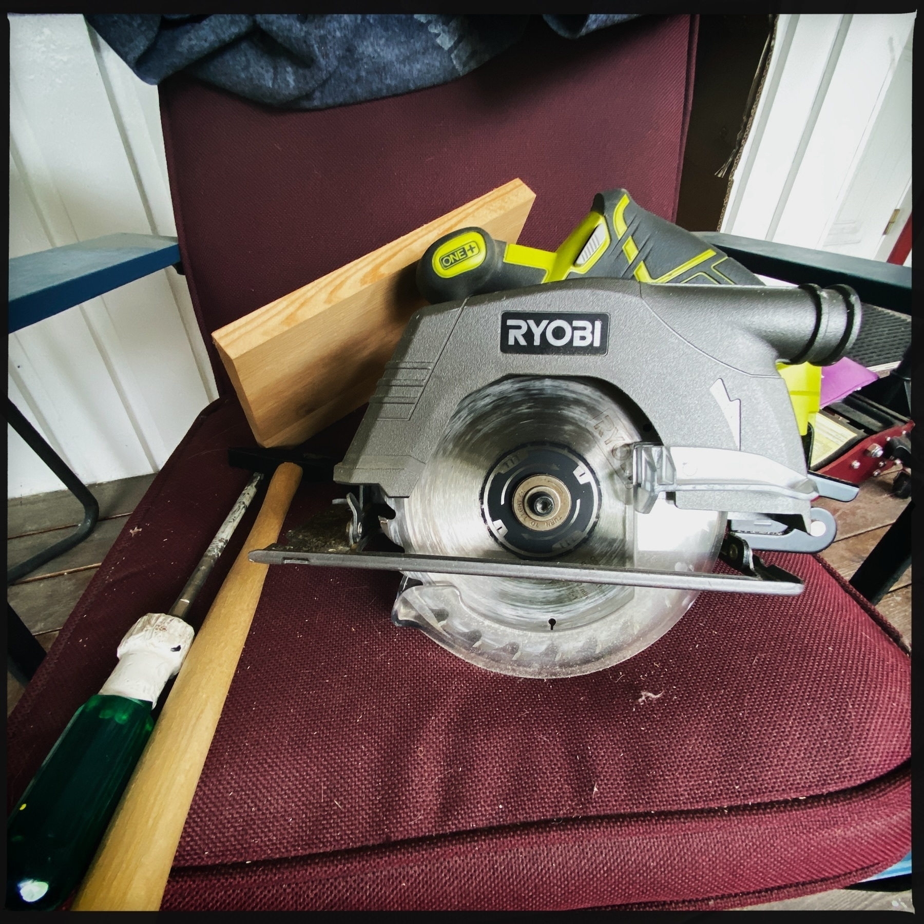 The image shows a Ryobi circular saw placed on a burgundy chair, alongside a chisel with a green handle and a piece of wood.