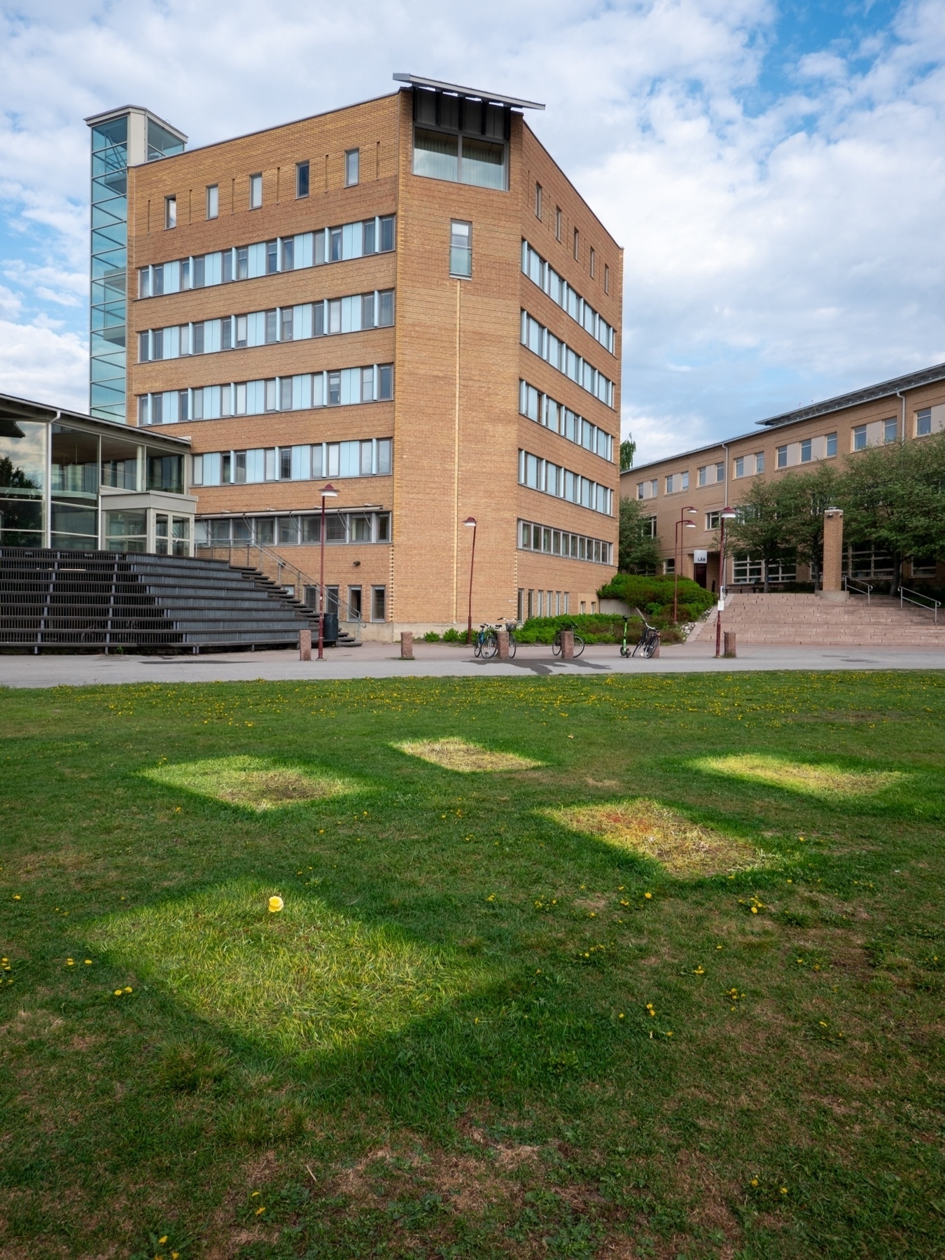 A multi-story brick building with large windows, an adjacent glass staircase tower, and another connected building in the background. The foreground features a grassy lawn with distinct square patches of light and some small yellow flowers. There are a few bicycles parked near the buildings
