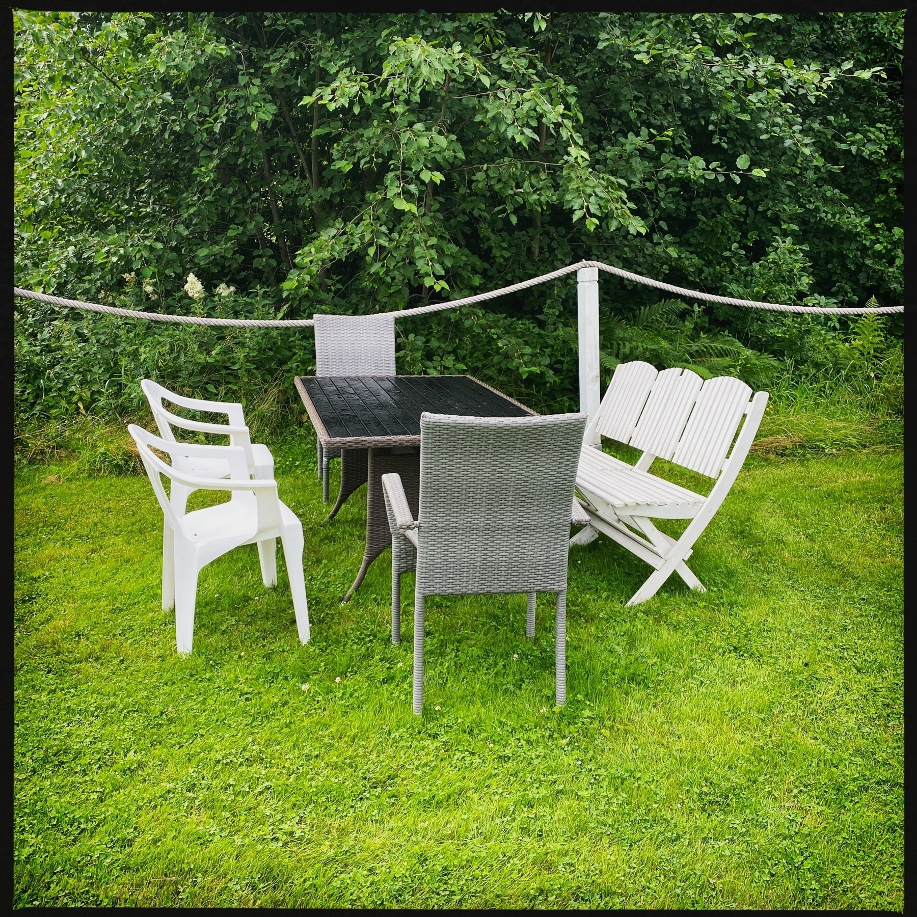 Outdoor patio setup with a rectangular black table, assorted chairs, and a white bench on a grass lawn. Forest greenery in the background and a white rope barrier.