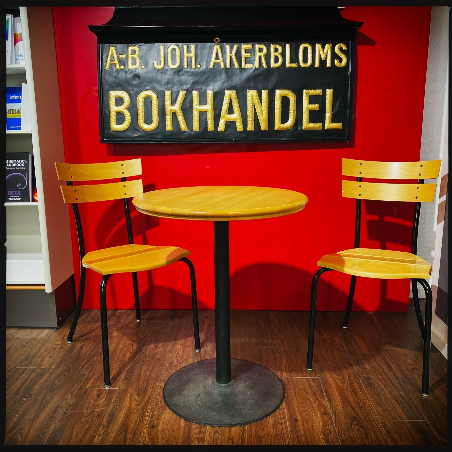 A small seating area featuring an orange round table and two matching chairs with metal legs is set against a bright red wall. A vintage sign in Swedish above the table reads “A.B. Joh. Åkerbloms Bokhandel” in bold yellow letters