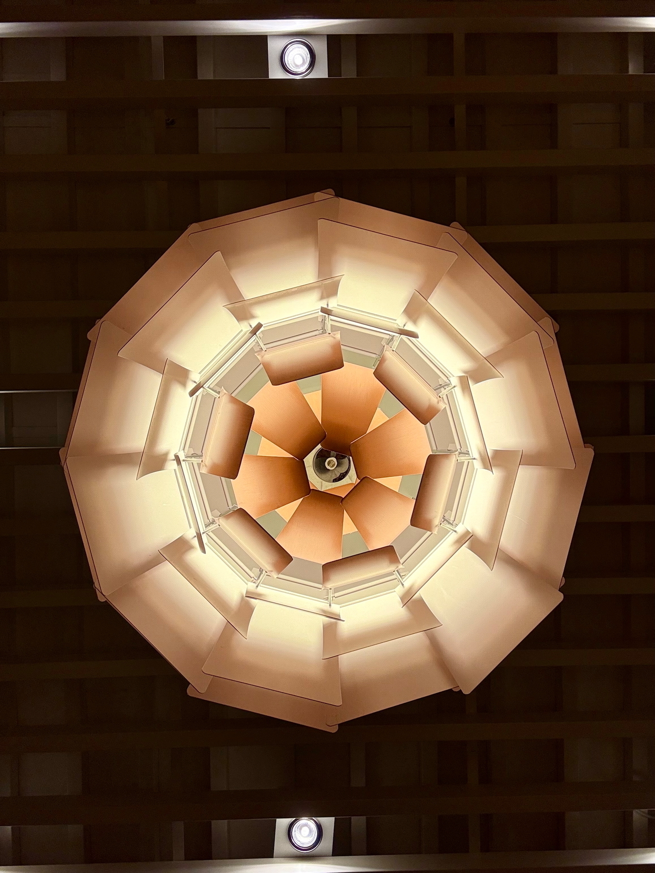 A geometric, abstract ceiling light fixture with layered panels radiates warm tones. The fixture is centered against a dark, grid-patterned ceiling.