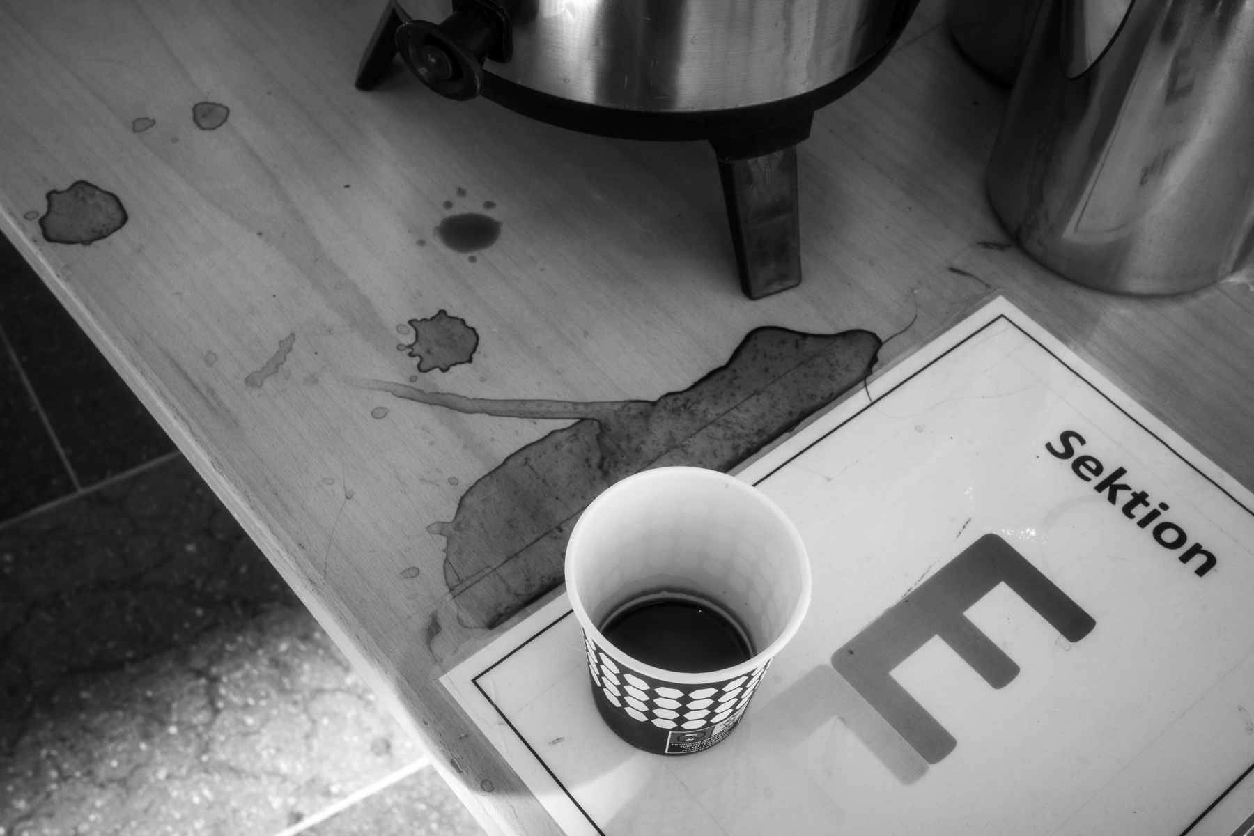 A black-and-white image showing a messy coffee station with a paper cup partially filled with dark liquid, likely coffee, placed on a table. There are various coffee stains and spills on the table. The corner of a sign labeled “Sektion E”