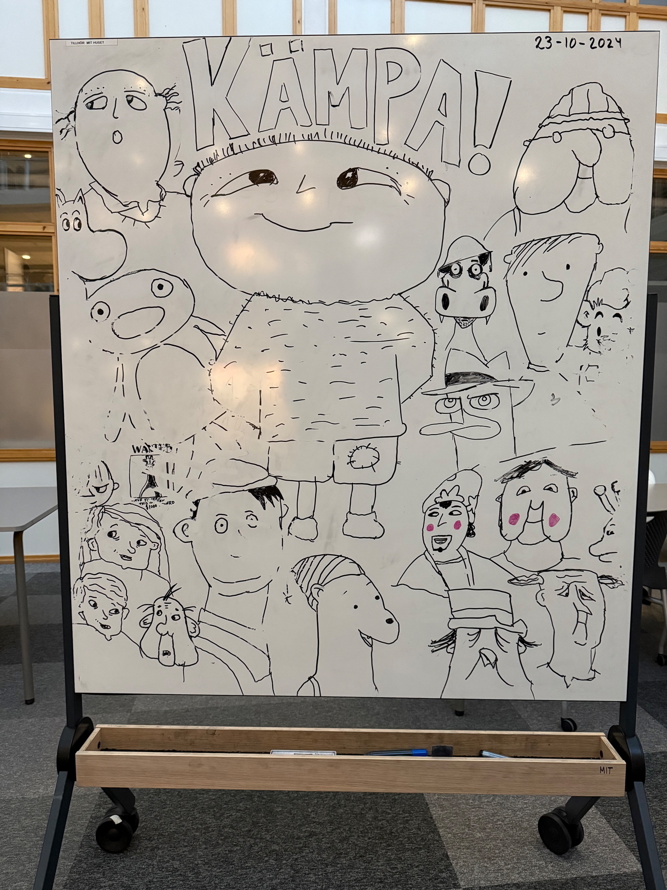 A whiteboard with a variety of cartoon-style characters drawn in black marker. At the top is the word “KÄMPA!” and the date “23-10-2024.” The characters have diverse expressions and designs, creating a lively and