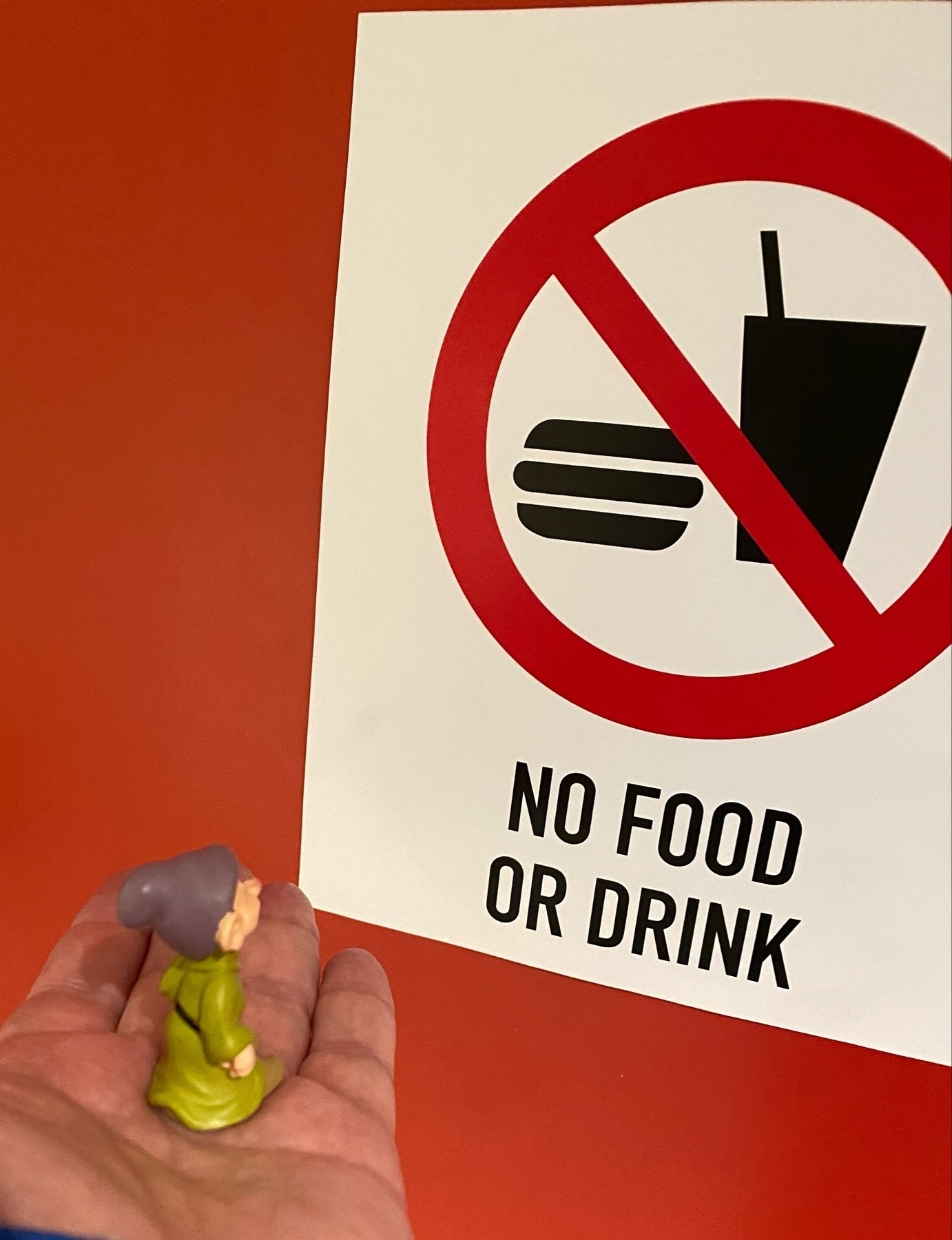 A person’s hand holding a small figurine in front of a sign that reads “NO FOOD OR DRINK” with a graphic symbol of no beverages and no burgers allowed.