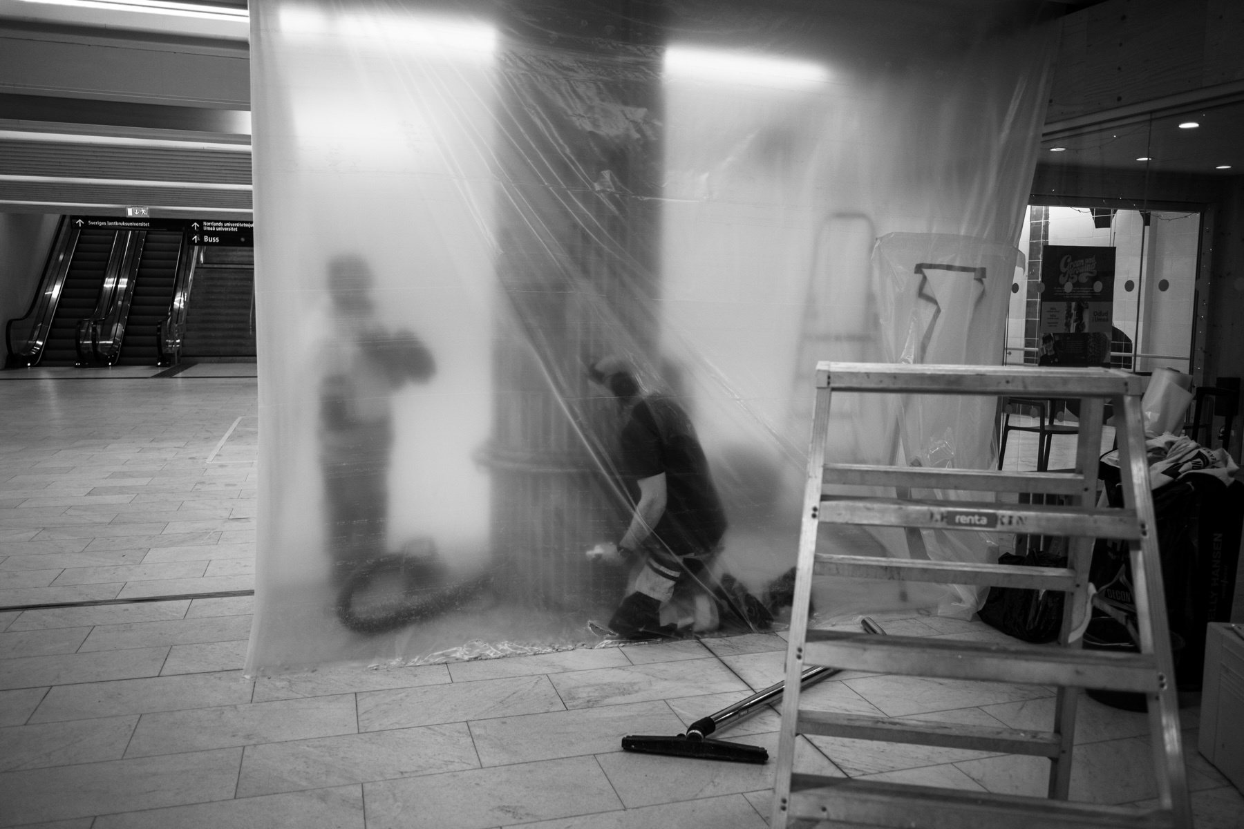 A dimly lit construction scene in a public space. Two workers are partially visible behind a translucent plastic sheet, engaged in work. A ladder, vacuum hose, and tools are visible in the foreground. Escalators and signage can be seen in the