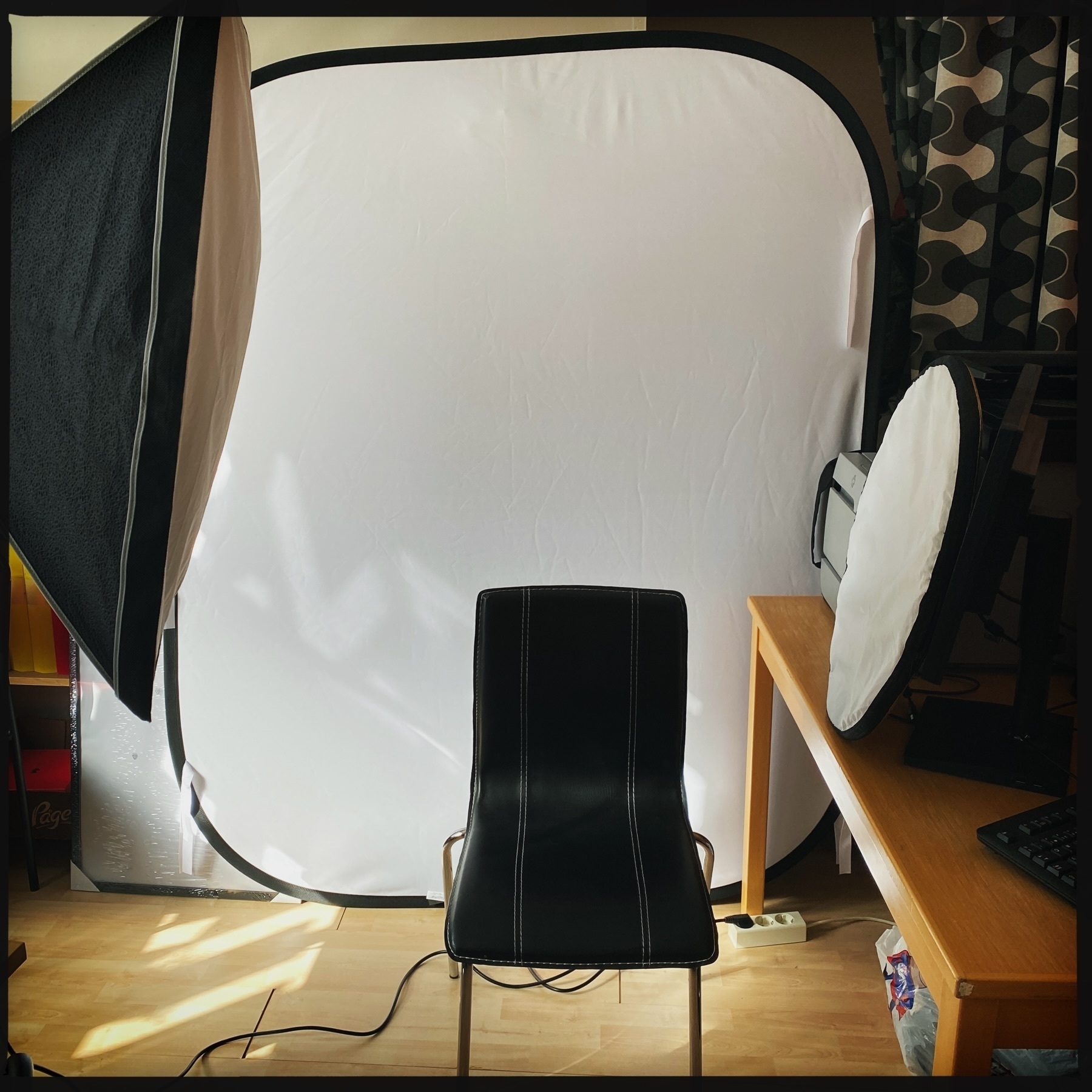 A minimalist home studio setup with a black chair centered in front of a large white backdrop. Surrounding the chair are a softbox light on the left, a desk on the right with a computer monitor and reflector, and some scattered cables on the wooden