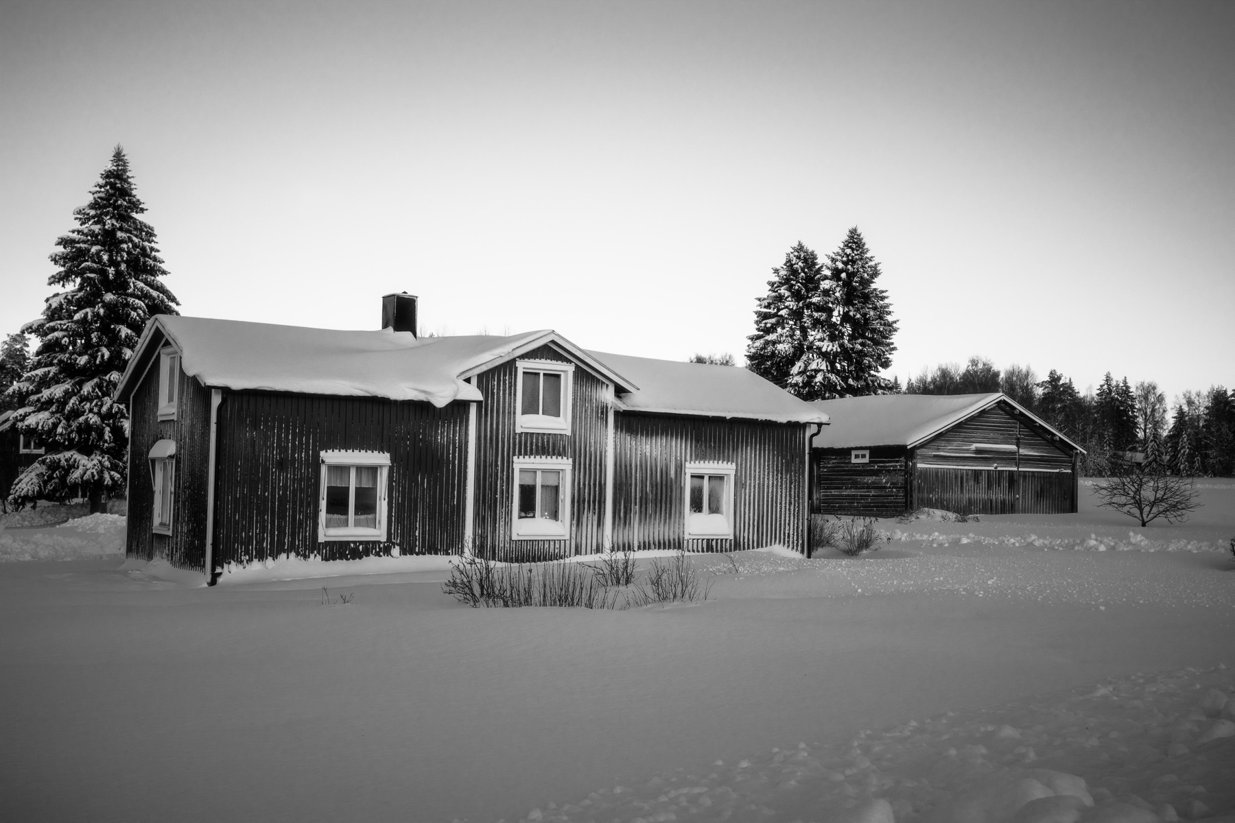 A snow-covered wooden cabin and an adjacent barn are set in a wintry landscape, surrounded by snow-laden trees under a clear sky.