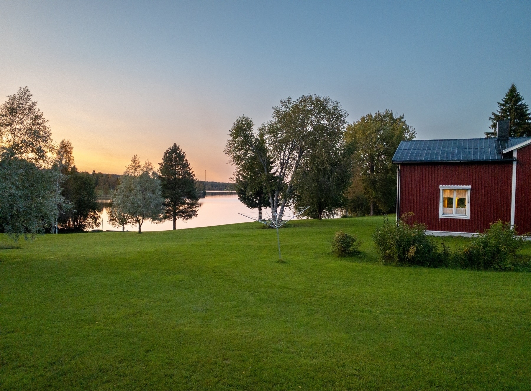 Red house with white trim, adjacent lawn, and lush green grass. Trees in the background surround a calm lake reflecting the evening sunset glow. Clear, open sky above.