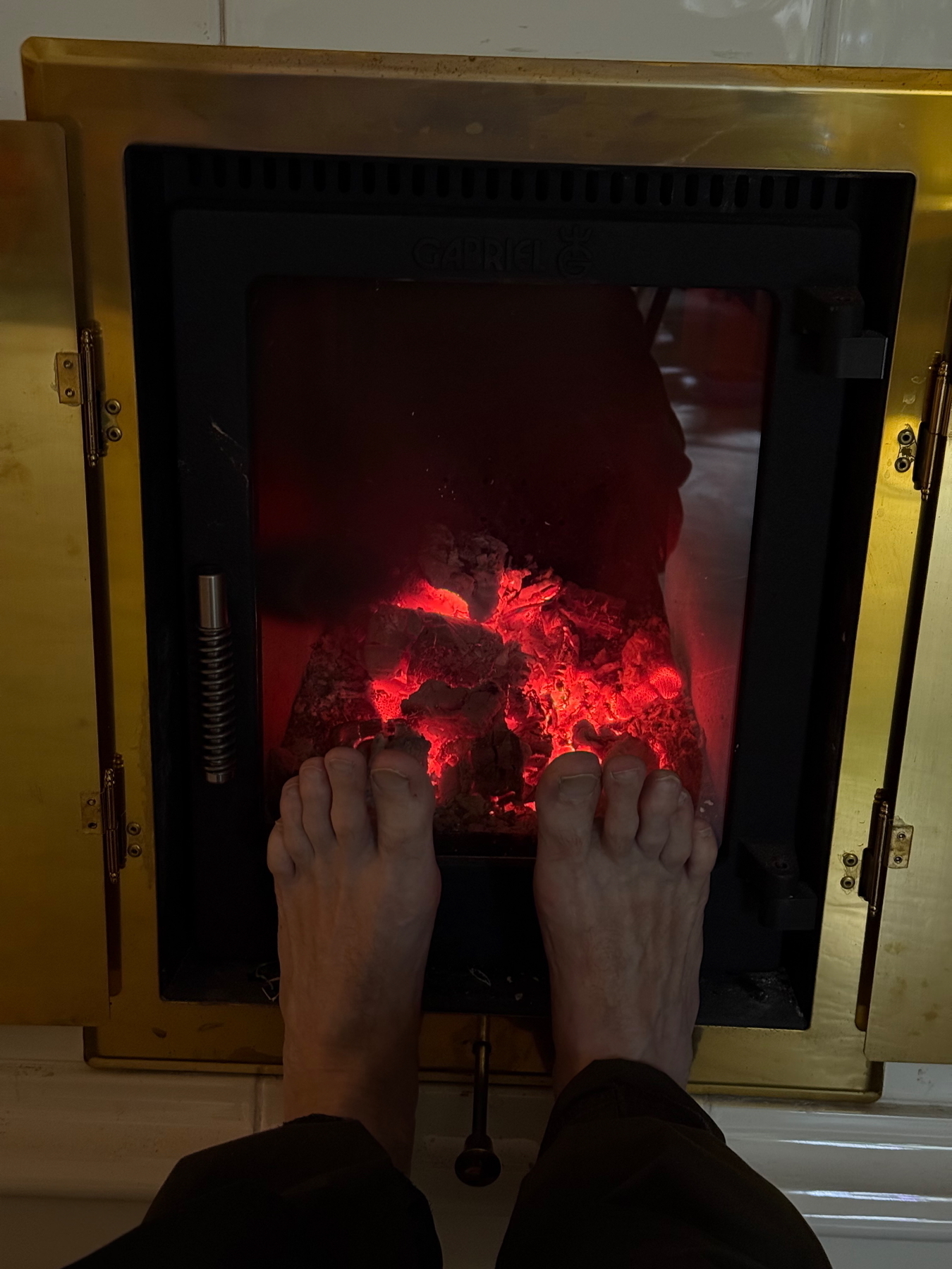 A pair of feet warming in front of a burning fireplace with glowing embers, enclosed by a brass-framed glass door.