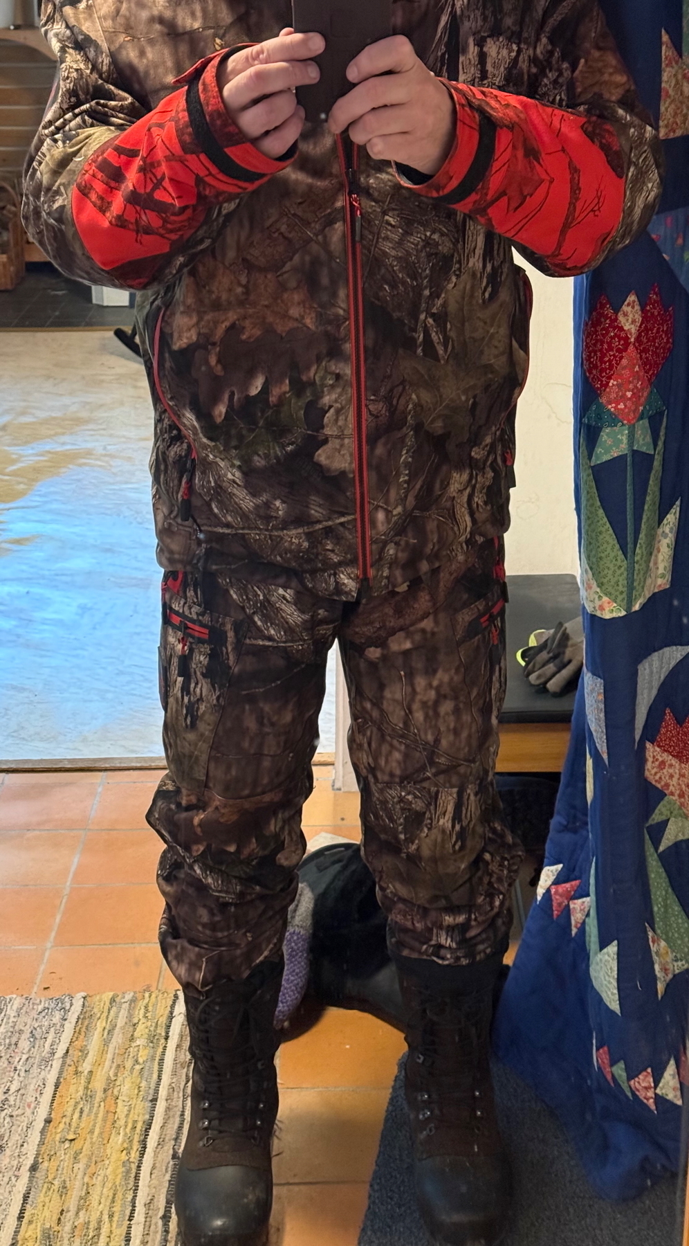 A person wearing a camouflage hunting outfit with orange accents and black boots, taking a mirror selfie. The background includes a quilt and a rug on the floor.