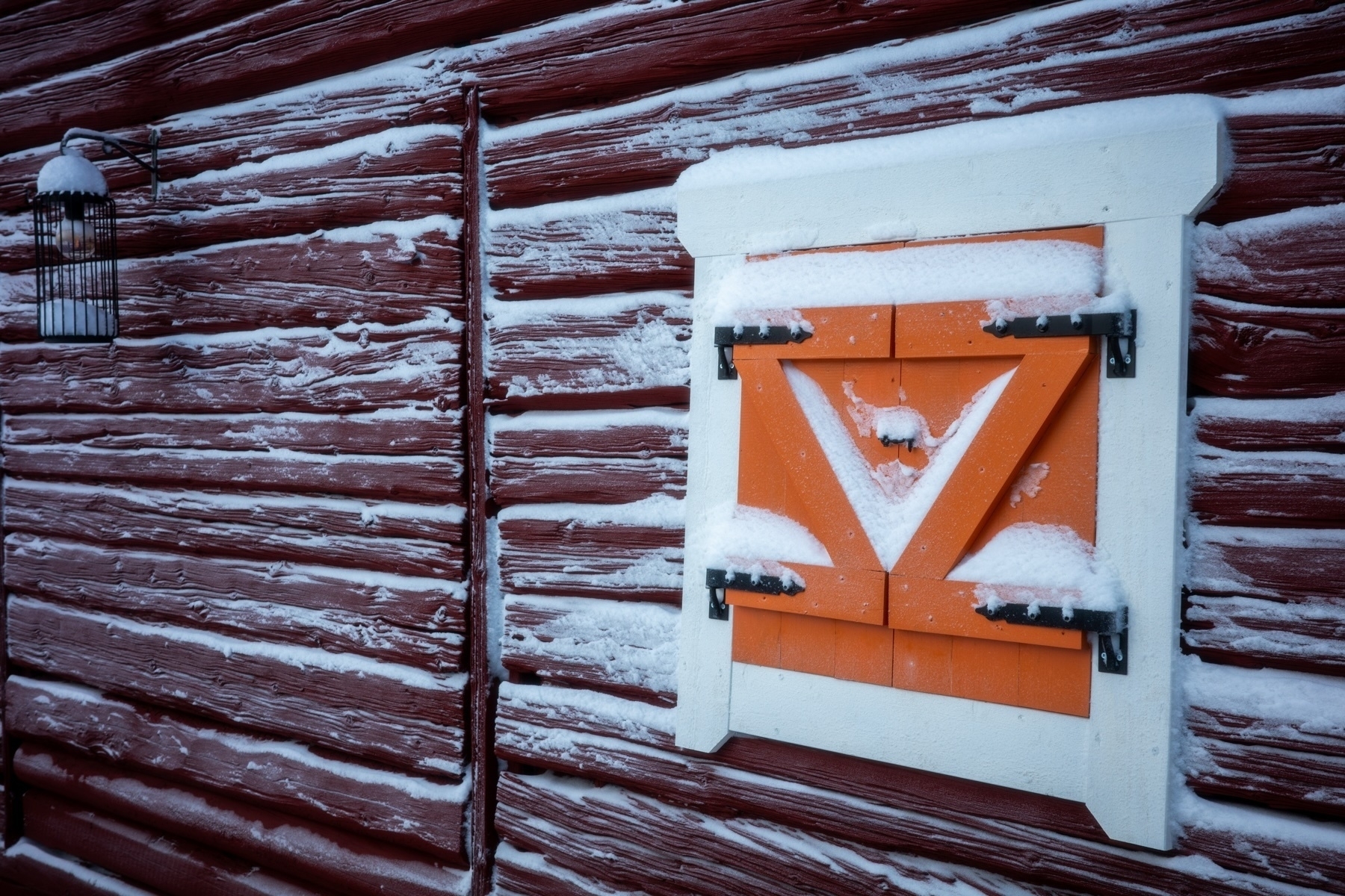 A snowy red wooden wall with a small orange window covered in snow. A black metal bracket and snow-covered lantern are visible.