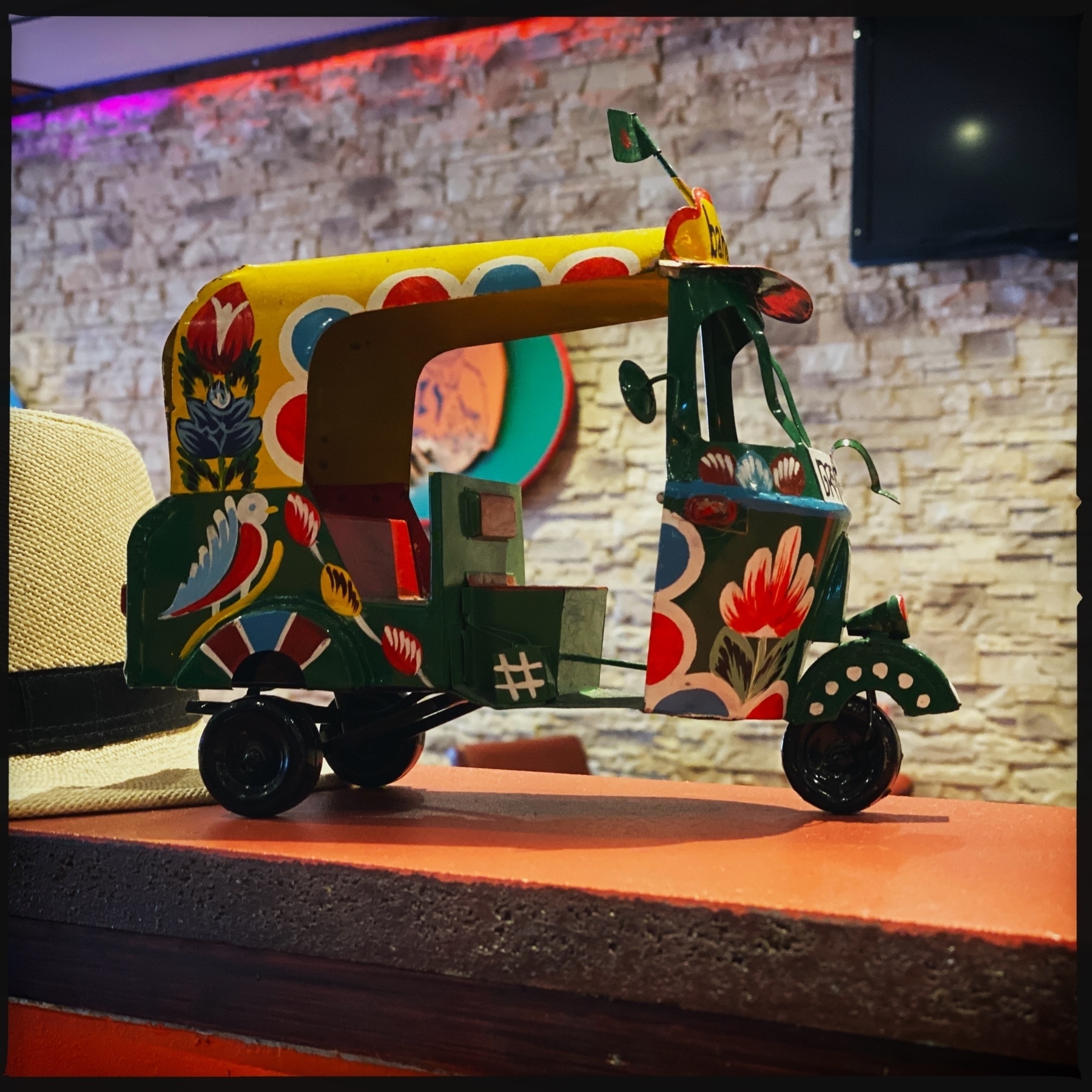 A brightly painted miniature rickshaw sits on a counter. The rickshaw features colorful floral designs and patterns. A cream-colored hat is partially visible to the left. The background includes a textured wall and some ambient lighting.