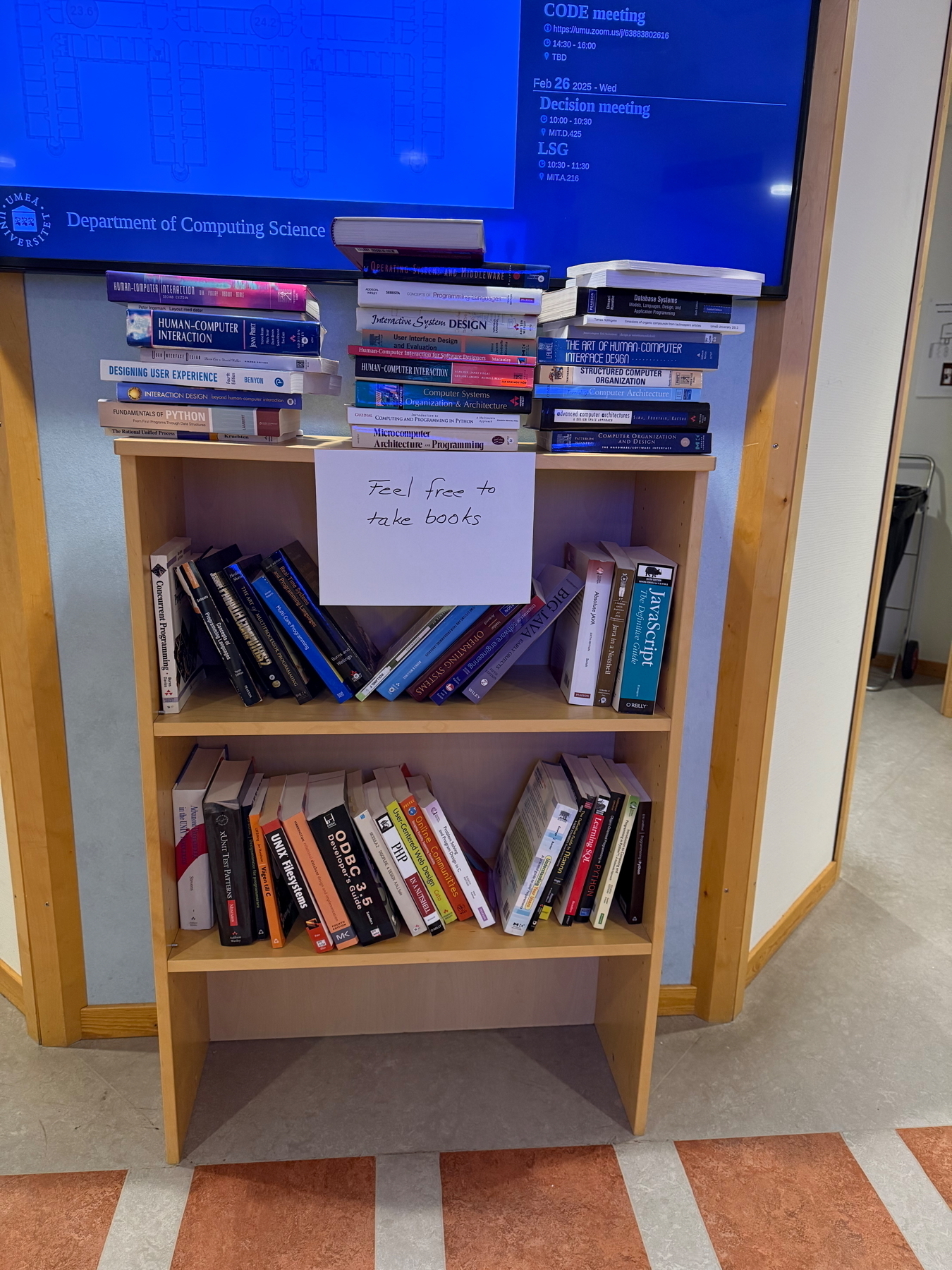 A wooden bookshelf filled with computer science and technology books is in a hallway. A sign on the top shelf reads “Feel free to take books.” There are stacks of books on top of the shelf as well. In the background, a digital screen displays