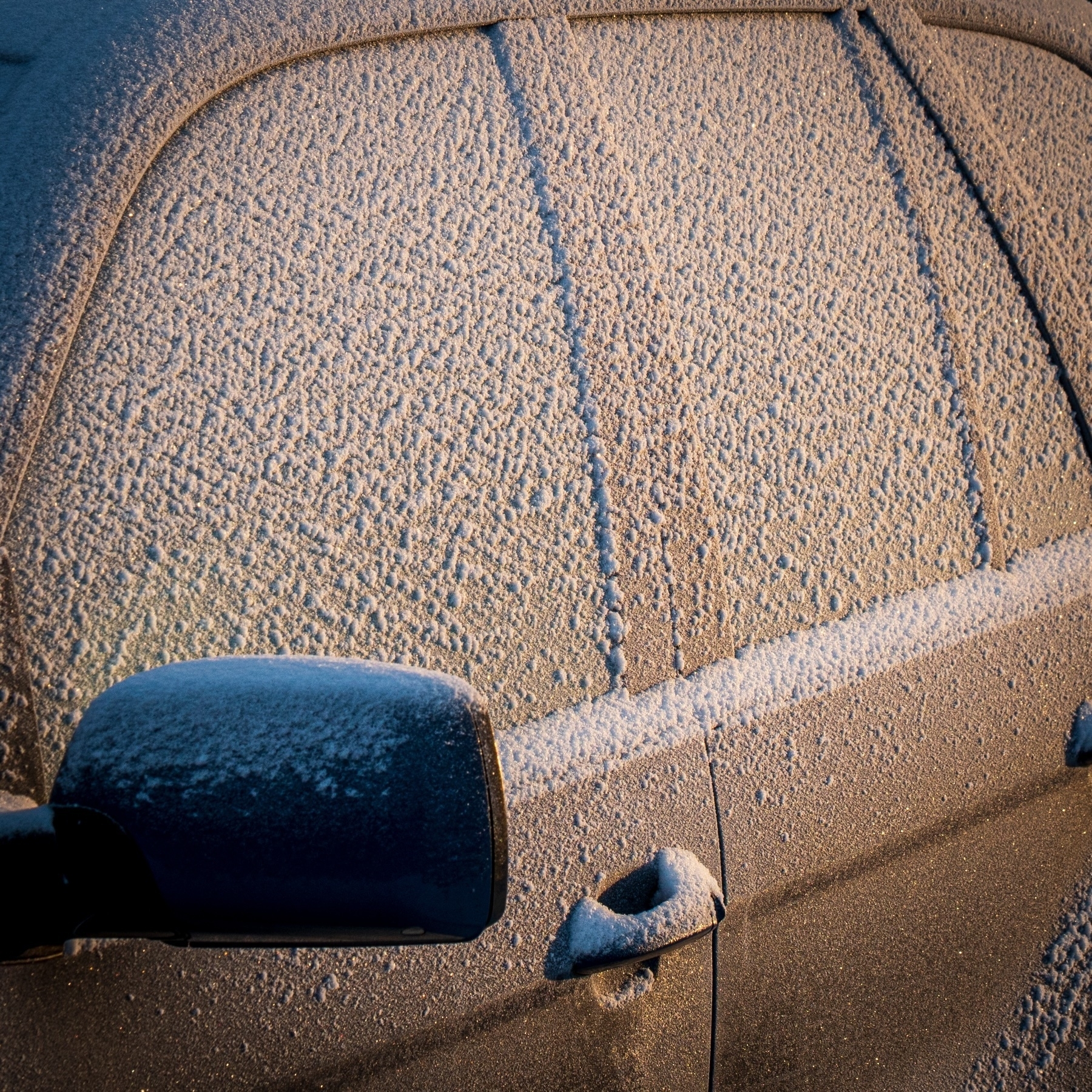 A car covered in frost, with ice crystals visible on the windows, side mirror, and door handle, illuminated by warm morning light..